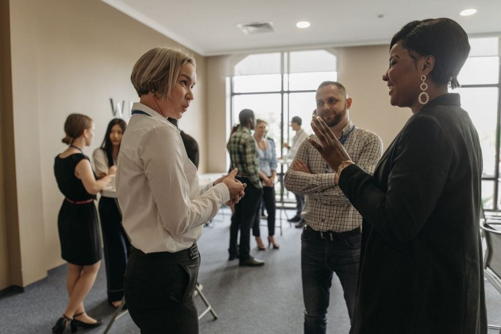 Profissionais conversando em pé durante um evento de networking, em um ambiente corporativo com outras pessoas reunidas ao fundo.