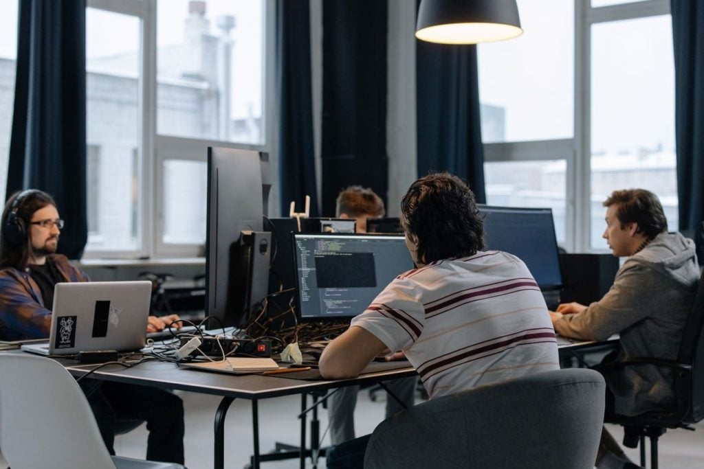 Equipe de pessoas desenvolvedoras programando em frente a monitores com código em um escritório, parte delas usando fones de ouvido.