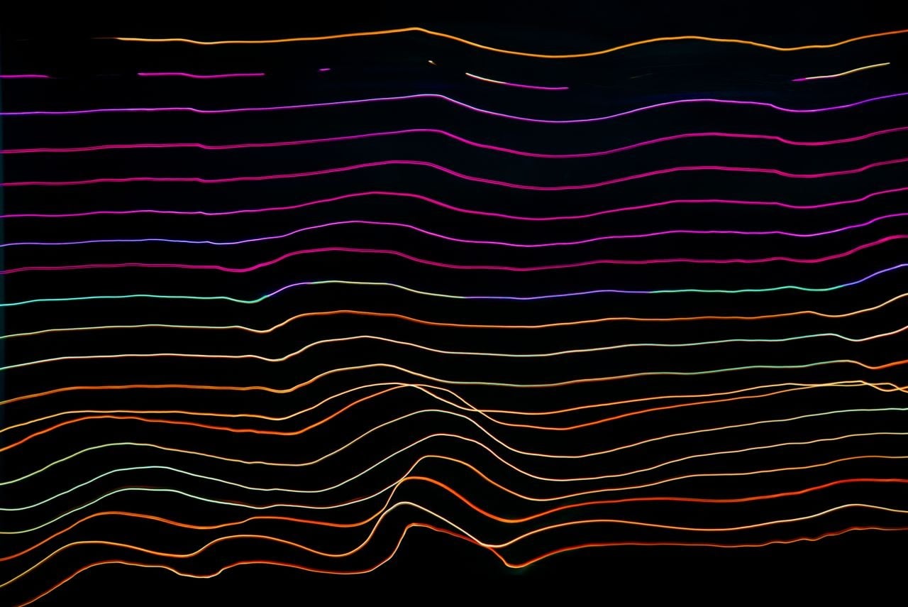 Linhas de luz coloridas formando ondas horizontais sobre fundo preto, remetendo a ondas sonoras de um áudio.