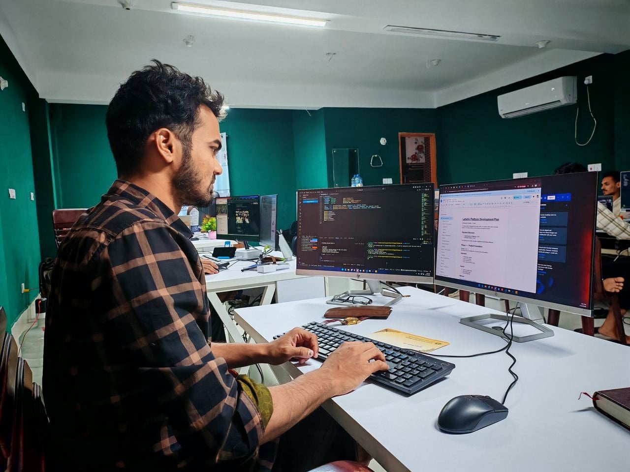 Alt-text: Pessoa programando em um escritório com dois monitores, um exibindo código e o outro um plano de desenvolvimento de plataforma.