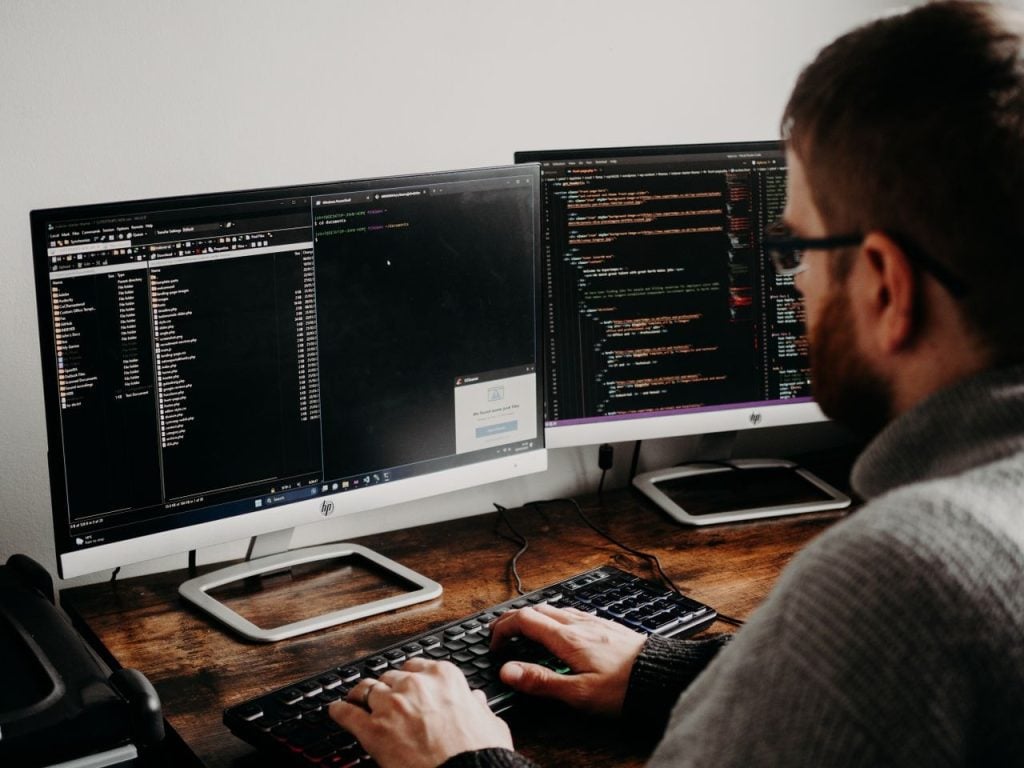 Pessoa trabalhando em dois monitores com terminal, gerenciador de arquivos e código-fonte abertos simultaneamente.
