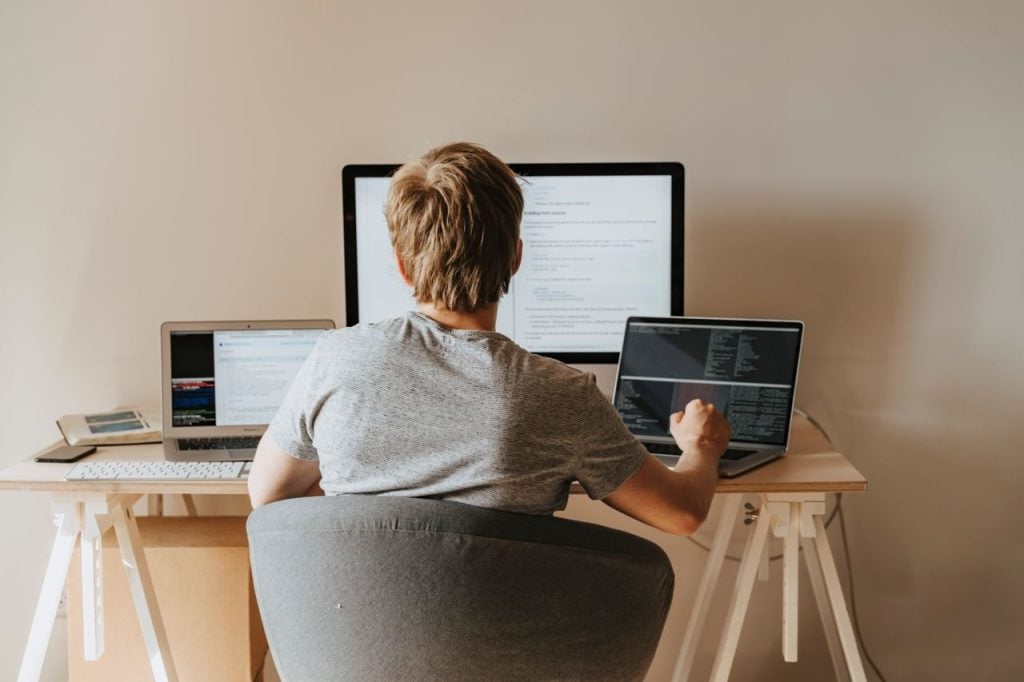 Pessoa vista de costas em uma mesa com três telas, consultando documentação técnica enquanto escreve código no notebook.