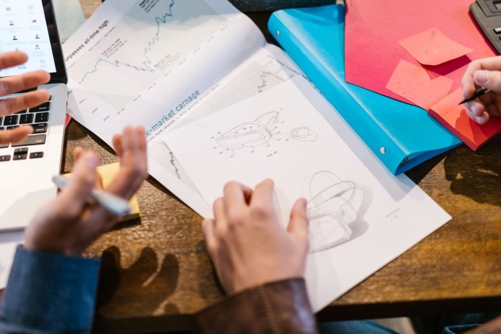 Duas pessoas analisando esboços de um produto sobre a mesa, ao lado de gráficos de mercado, notebook e anotações coloridas.