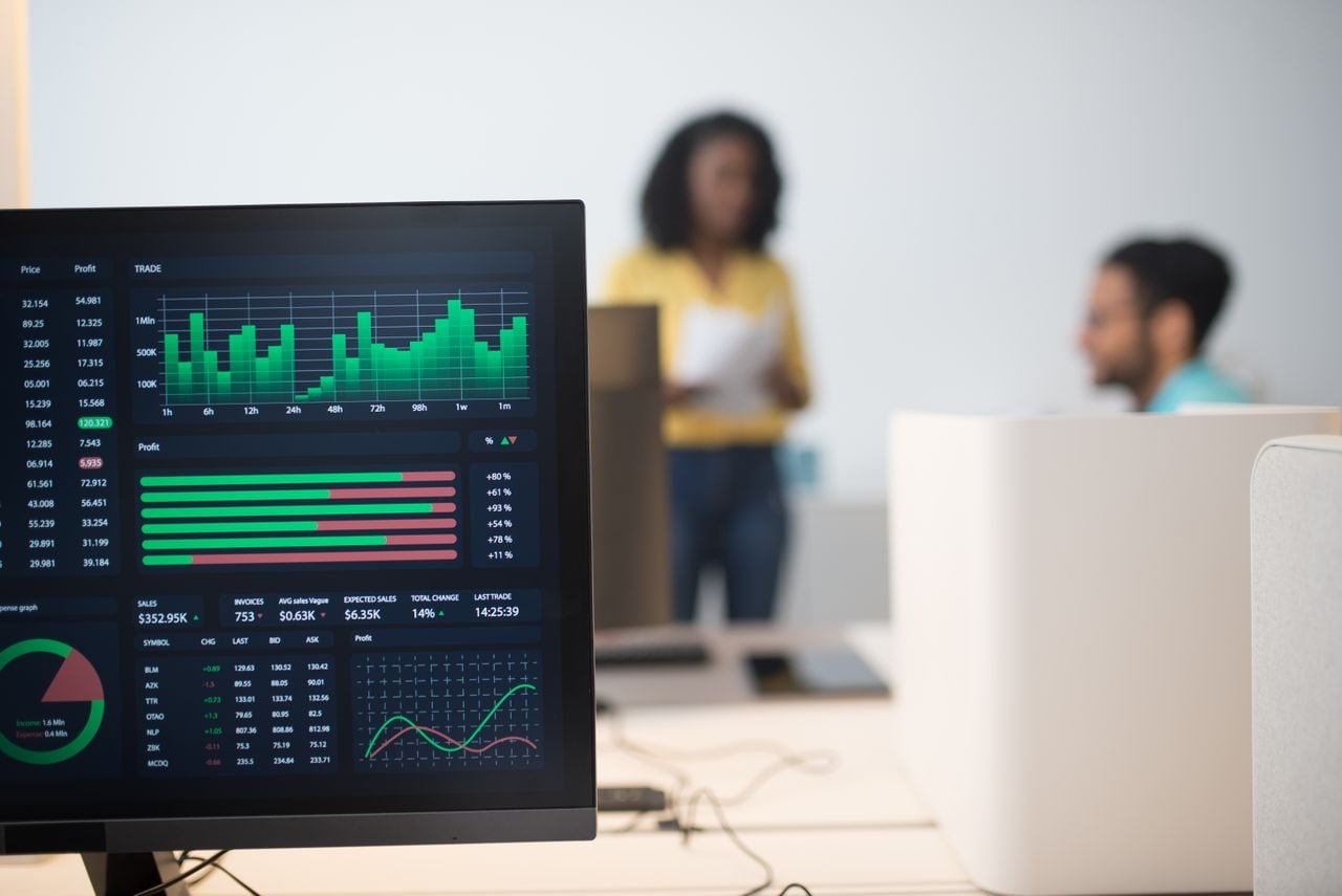 Monitor exibindo um painel com gráficos, tabelas e indicadores de desempenho, com duas pessoas ao fundo em um escritório.