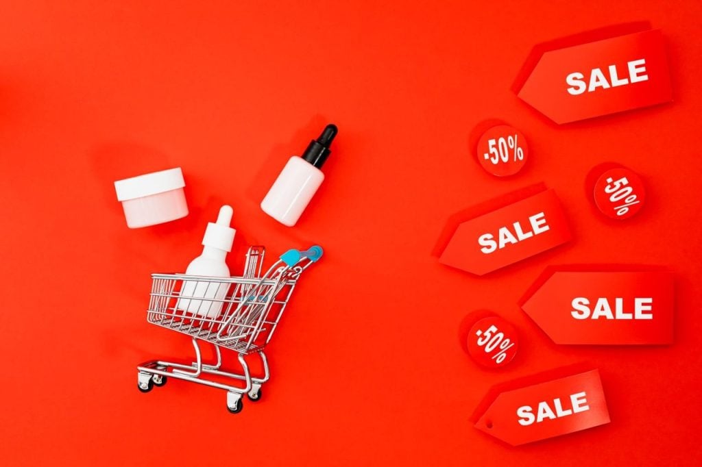 Um mini carrinho de compras de supermercado prateado e azul no canto inferior esquerdo de um fundo vermelho vibrante, cercado por produtos de beleza (creme e séruns) e diversas tags vermelhas de promoção escritas 'SALE' e '-50%'.