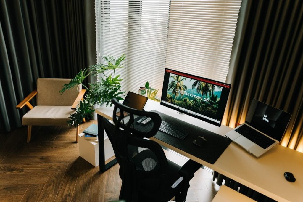 Ambiente de home office moderno e organizado, com uma mesa contendo um computador desktop principal, um notebook aberto e um tablet, além de uma planta grande e uma poltrona, iluminado por luz natural vinda de uma janela com persianas.