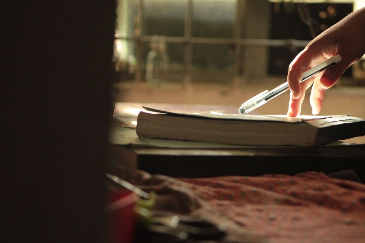 Mão segurando uma caneta sobre um livro aberto em uma mesa junto à janela, com luz natural iluminando a página.