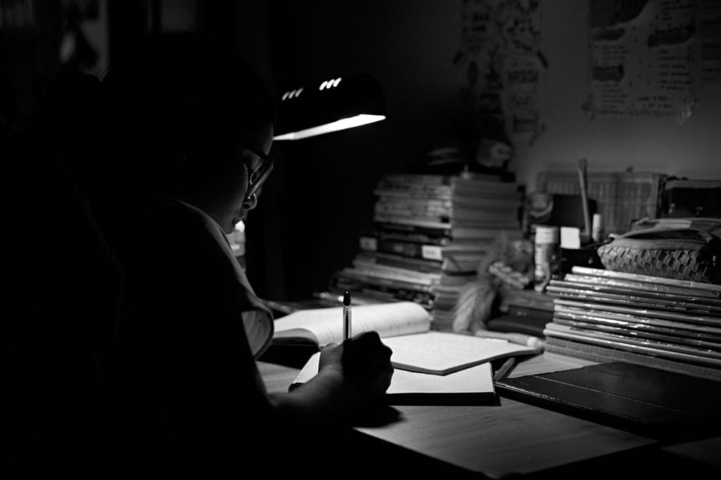 Pessoa de óculos escrevendo em um caderno sob a luz de uma luminária de mesa, cercada por pilhas de livros, em preto e branco.